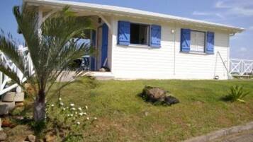 A single-story white house with blue shutters, a porch, and a small garden with a palm tree.
