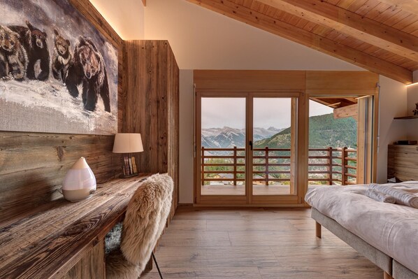A bedroom with a wooden headboard, a painting, a lamp, a fur throw, a bed, and a view of mountains through a glass door.