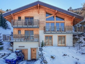 A two-story wooden chalet with a balcony on the upper floor, a garage with a car parked inside, and a stone wall in front. - Chalet Kudu - Jacuzzi - Sauna - Views - La Tzoumaz, 4 Vallees (La Tzoumaz)