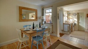 Un coin repas avec une table en bois, des chaises et un miroir au mur. Il y a une fenêtre avec des rideaux et une vue sur l’extérieur.