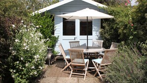 Une terrasse avec une table et des chaises sous un parasol blanc, entourée de verdure et d’un petit abri de jardin.