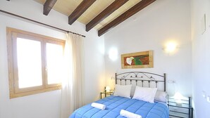 A bedroom with a bed, a window with curtains, a painting on the wall, and a nightstand with a lamp.