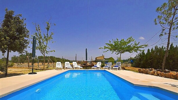 A rectangular swimming pool with clear blue water, surrounded by white lounge chairs and a stone wall.