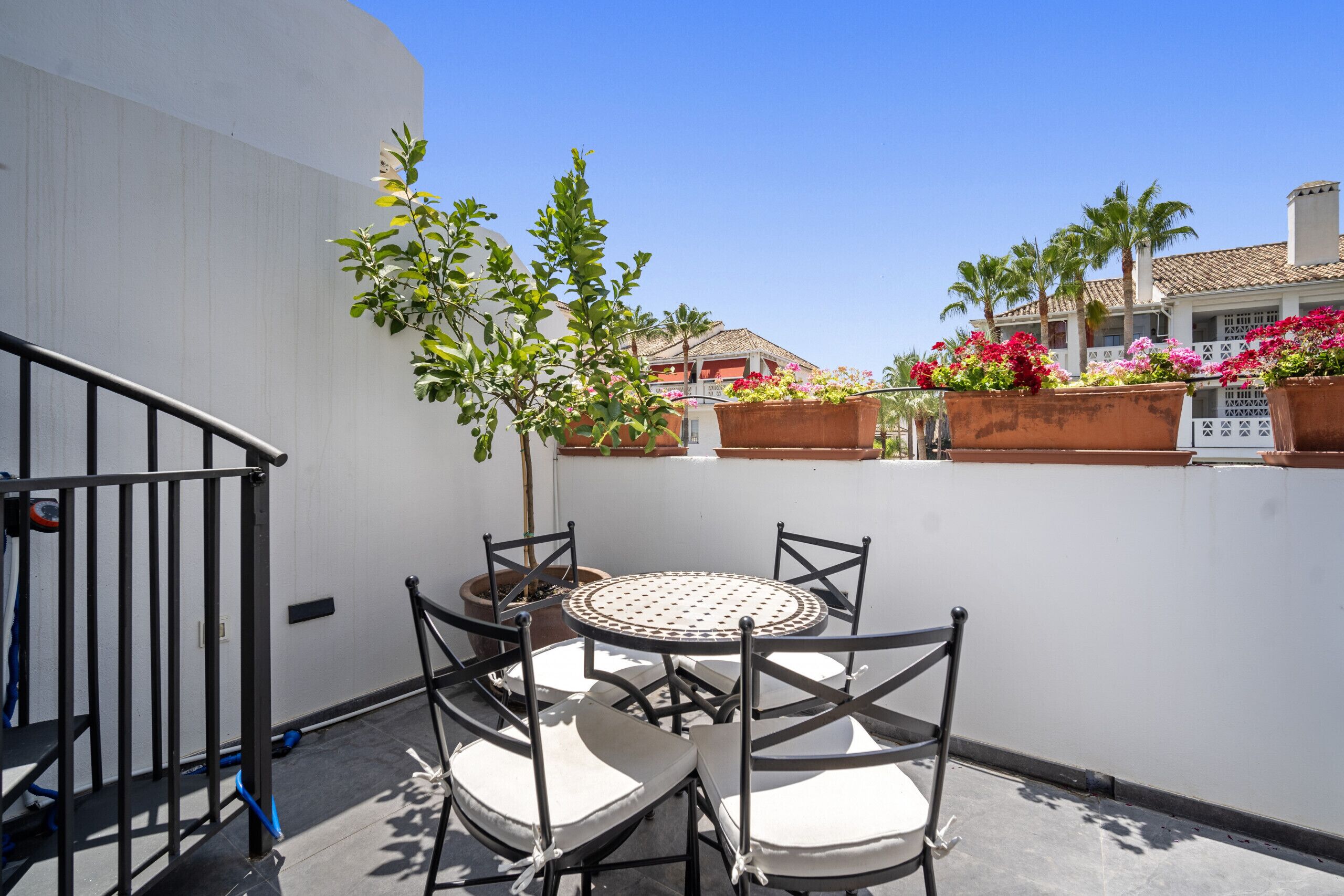 A balcony with a round table and four chairs, surrounded by potted plants and flowers.