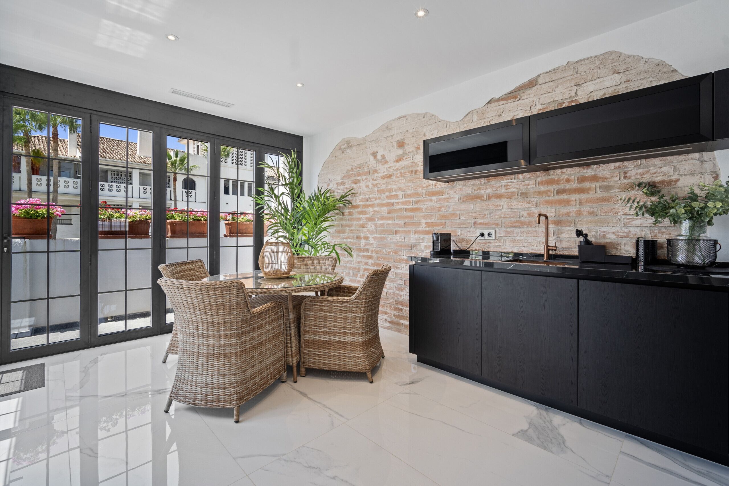 A modern kitchen with a brick wall, black cabinets, and a large window overlooking a courtyard with plants.