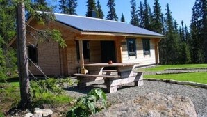 Una cabaña de madera con un porche cubierto y una mesa de picnic, rodeada de un área de grava y árboles.