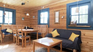 Interior de una cabaña de madera con una mesa de comedor, sillas, un sofá y una ventana con vistas a árboles.
