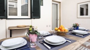 Une table à manger dressée avec des assiettes, des verres et des couverts, un panier de fruits et une plante en pot.