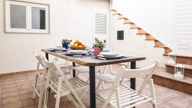 Un coin repas avec une table dressée pour quatre personnes, des chaises blanches, un saladier de fruits et une plante en pot.