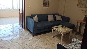 A living room with a blue sofa, a small white table, and a door leading to a balcony. - Floor apartment with superb roof terrace, next to Almirida sandy beach   (Chania)