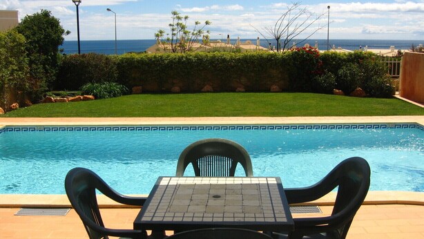 A pool with a view of the ocean, surrounded by greenery and a tiled deck with chairs.