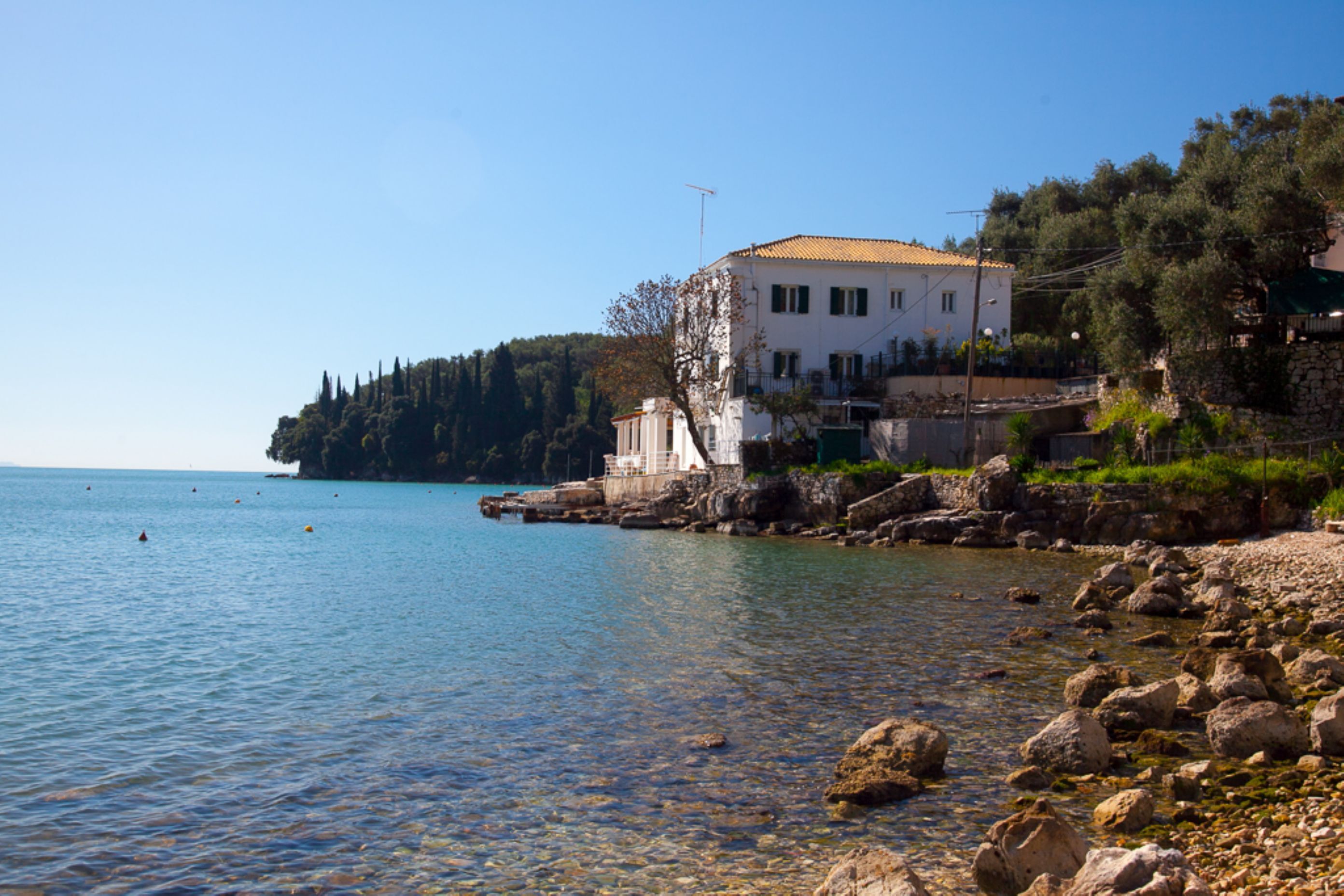 A coastal building with a tiled roof, situated on a rocky shoreline with clear blue water.