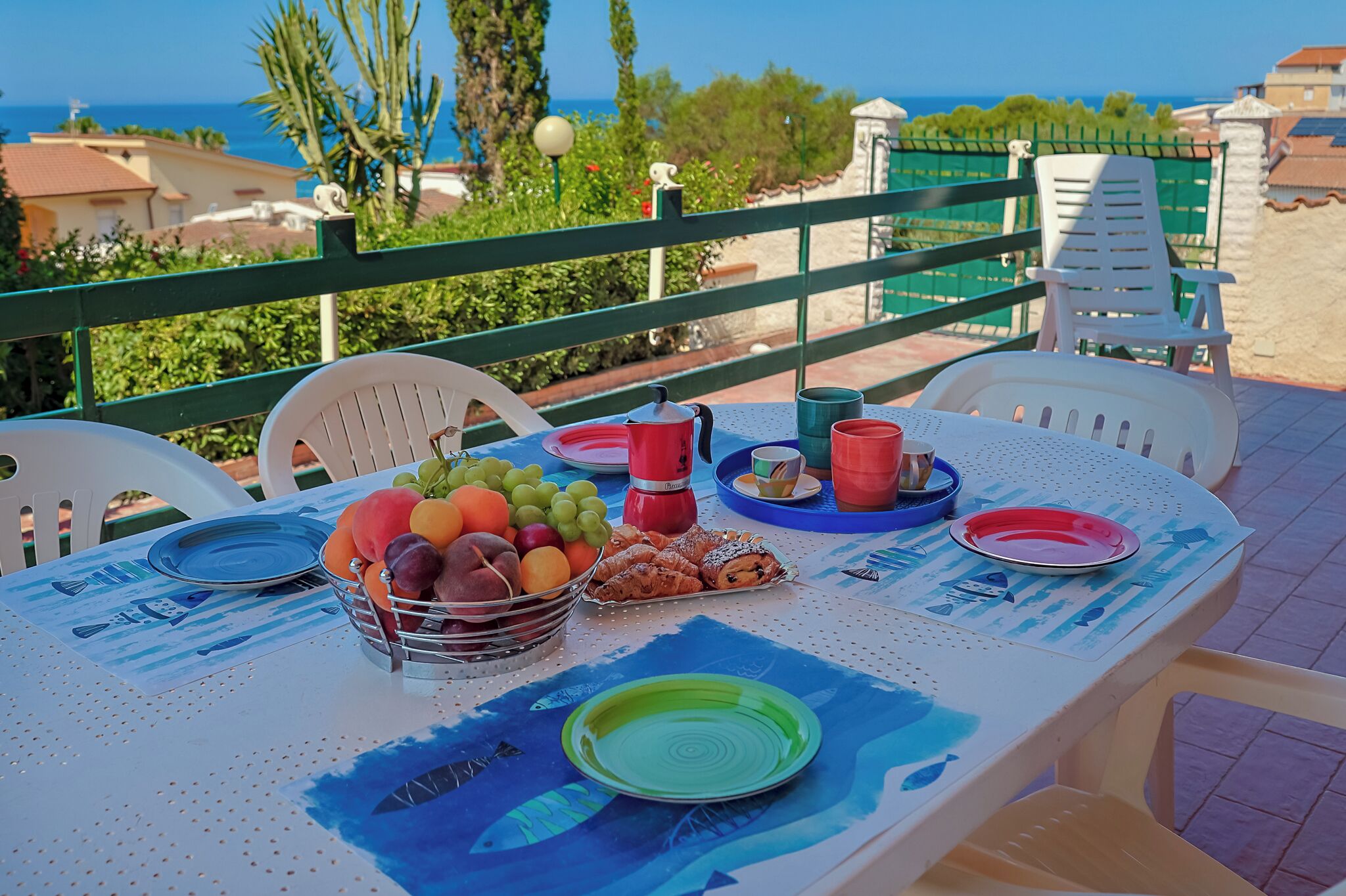 Ein Tisch mit Tellern, Tassen und einer Obstschale auf einem Balkon mit Blick auf das Meer.