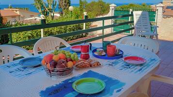 Ein Tisch mit Tellern, Tassen und einer Obstschale auf einem Balkon mit Blick auf das Meer.