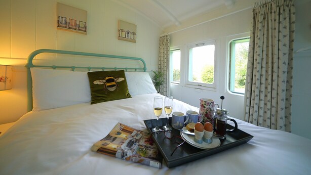 A neatly made bed with a tray of breakfast items, including toast, eggs, and coffee, in a room with a window and curtains. - Charming Seaside Home, Full of Character (Old railway carriages) Pet Friendly,  (Chichester)