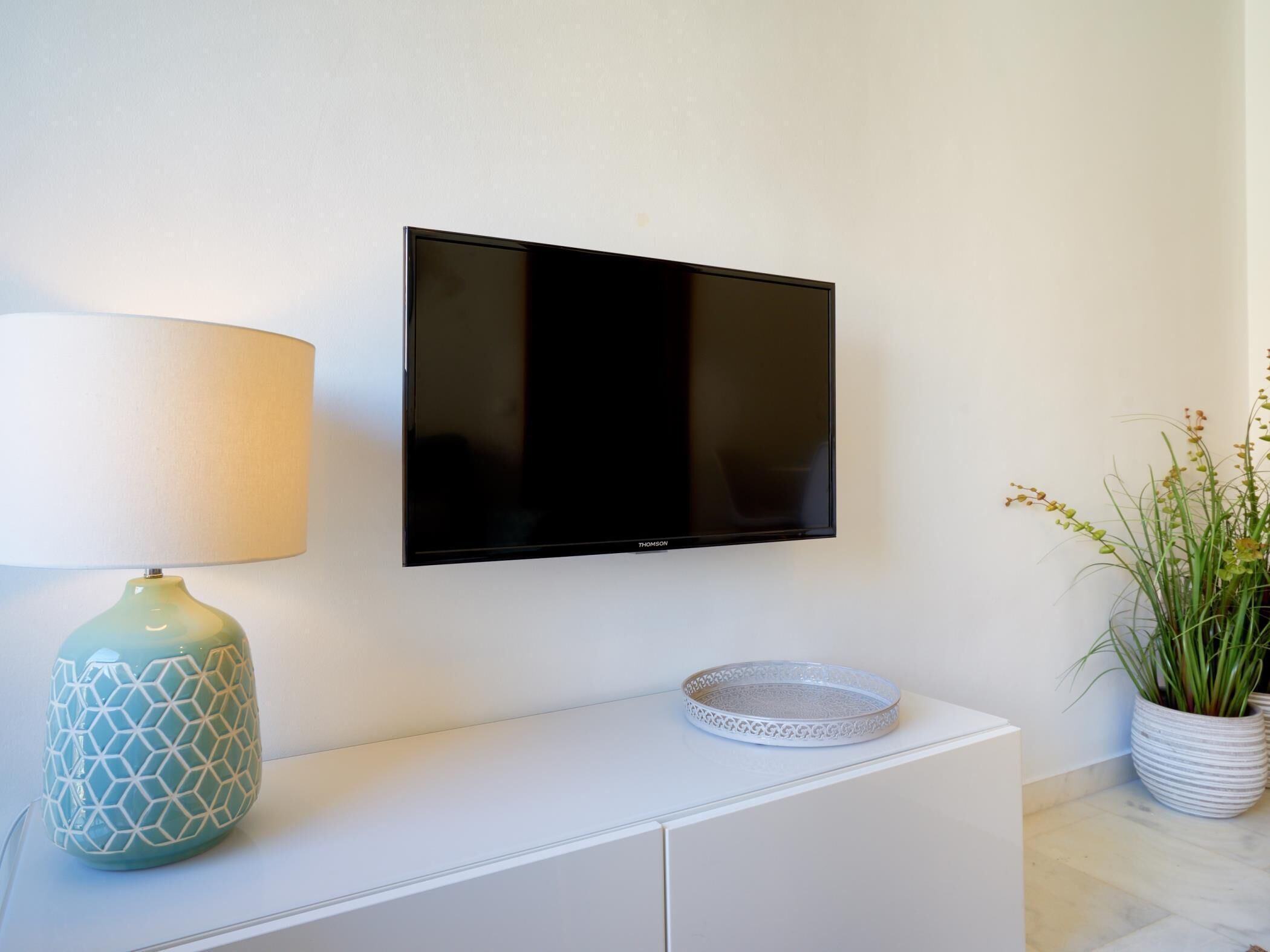 A flat-screen TV mounted on a white wall, a decorative lamp, and a potted plant beside a white cabinet.