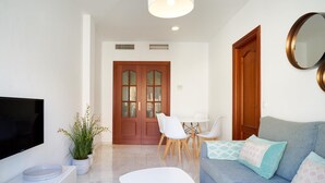 Una sala de estar moderna con un televisor de pantalla plana, un sofá con cojines estampados, un comedor con una mesa y sillas blancas, y una puerta de madera con paneles de vidrio.