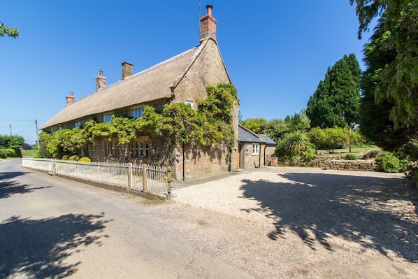 A stone house with a thatched roof, surrounded by a gravel driveway and lush greenery.