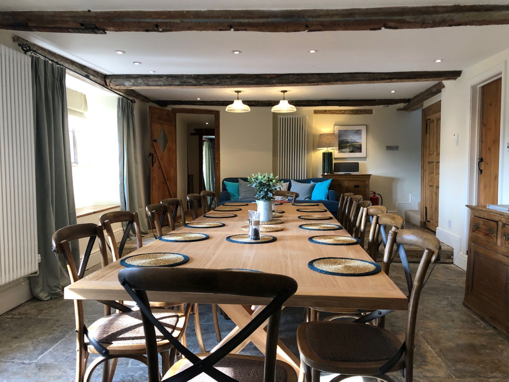 A dining room with a long wooden table set for 12, surrounded by wooden chairs, with a rustic ceiling and a view into the living area.
