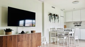 Een moderne keuken met witte kasten, een eettafel met stoelen, een aan de muur bevestigde televisie en een potplant.