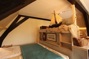 Een gezellige slaapkamer op zolder met een schuine kap, een bed met een blauwgroene sprei, een klein model zeilboot en een rieten mand.