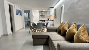 Un salon moderne avec un canapé d’angle gris, une table à manger avec des chaises, et une cuisine en arrière-plan.