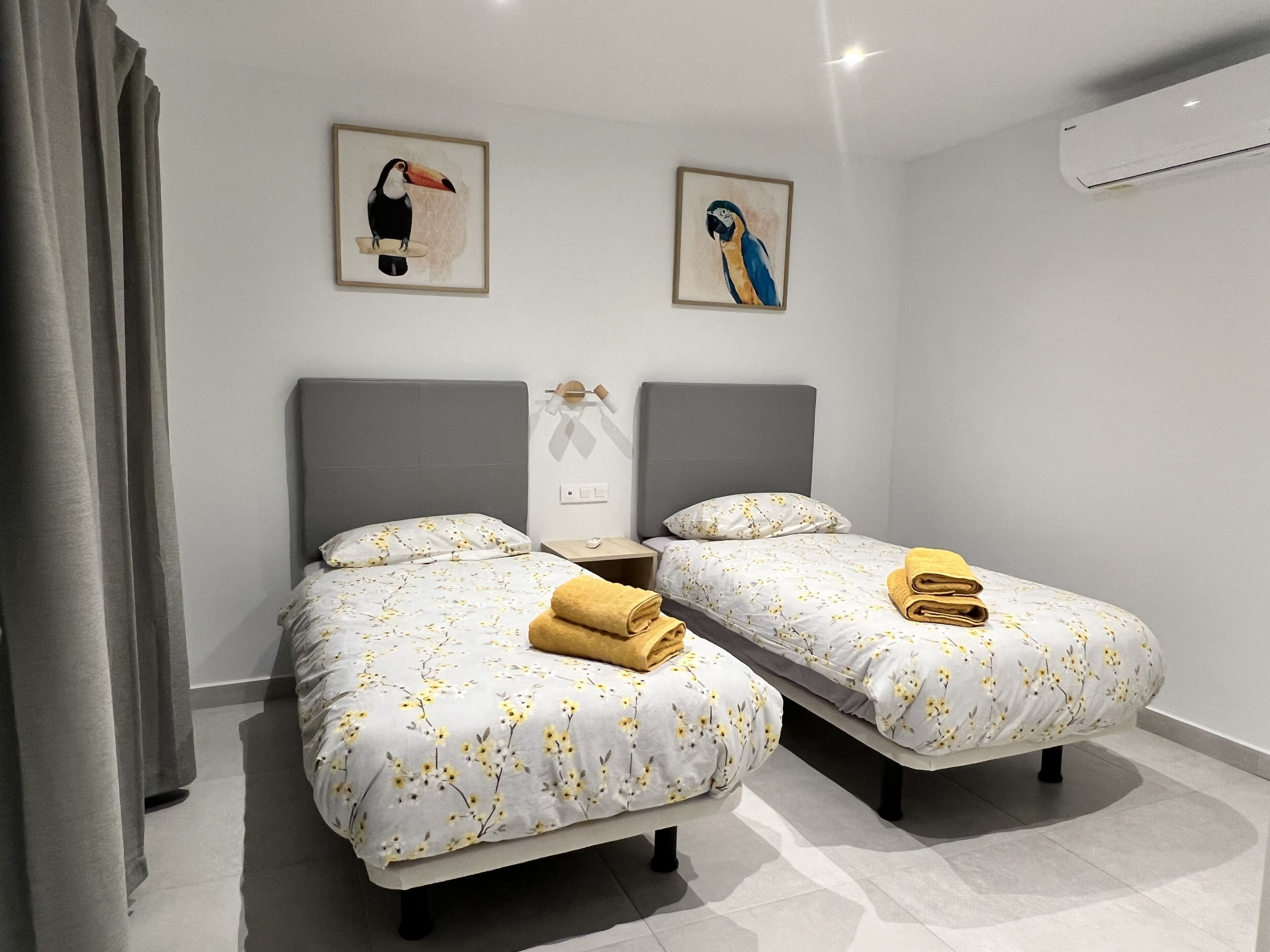 Une chambre d’hôtel avec deux lits simples, chacun recouvert d’une courtepointe à motifs floraux et agrémenté d’une serviette pliée. Deux tableaux représentant des oiseaux, encadrés, sont accrochés au mur au-dessus des lits.