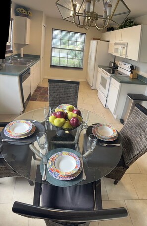 A dining area with a glass table, chairs, and a bowl of fruit. There is a kitchen in the background with white cabinets and appliances.