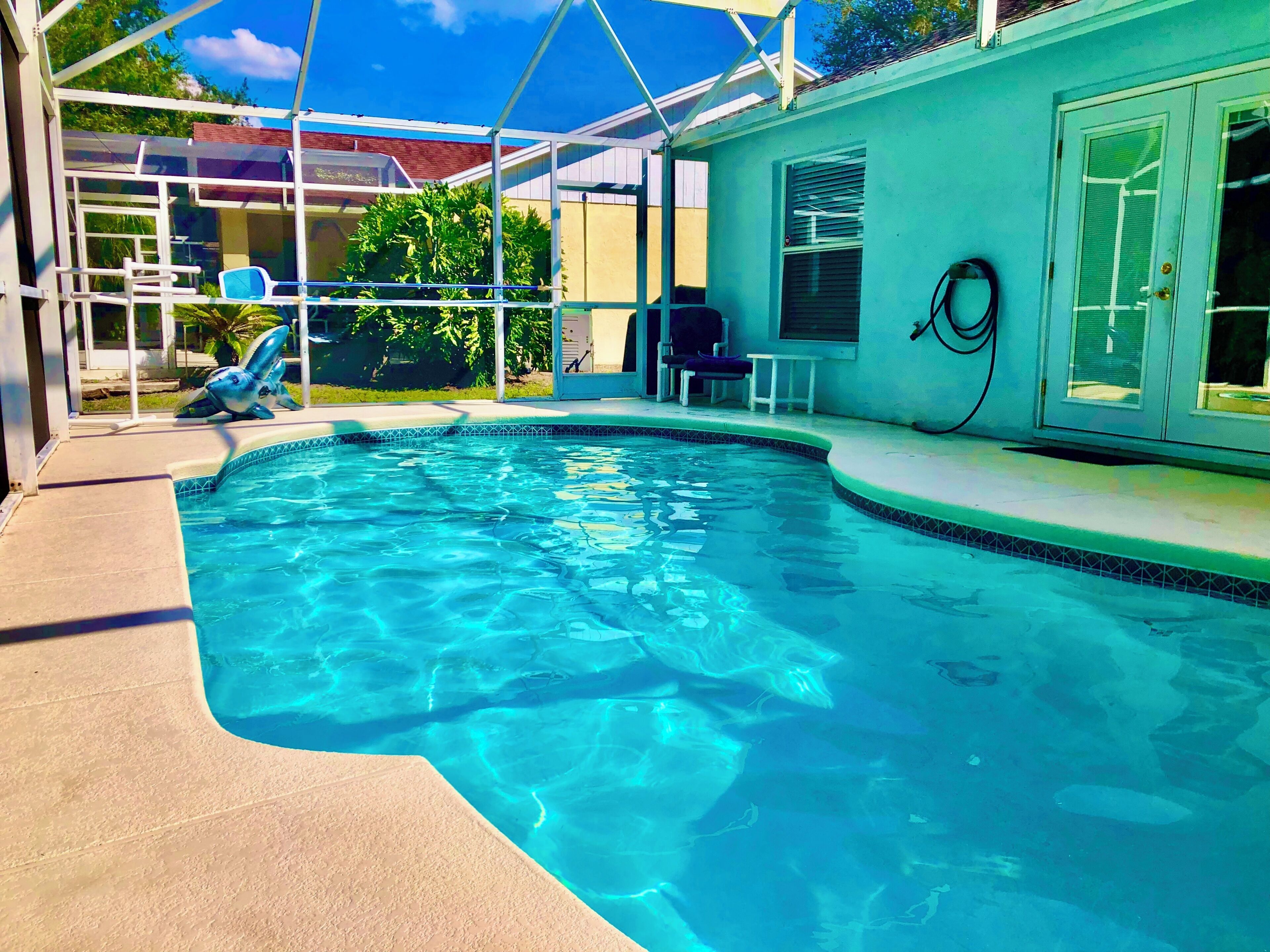 A clear swimming pool with a tiled edge, a covered seating area with chairs, and a hose on the wall.