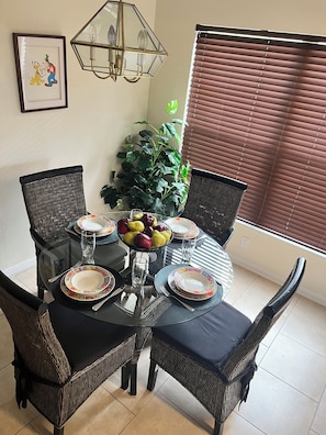 A dining area with a round table set for four, surrounded by wicker chairs. A large potted plant is in the background, and a framed picture hangs on the wall.