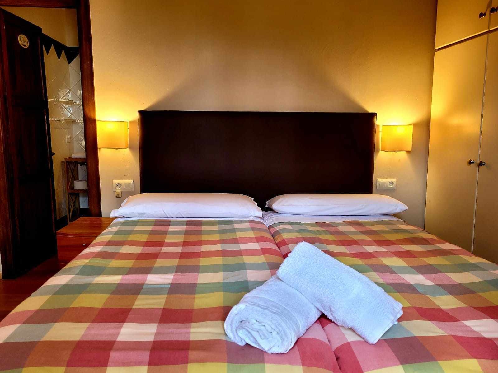 Un lit double avec une couvre-lit à carreaux colorés et deux oreillers blancs. Deux lampes murales sont fixées de chaque côté du lit.