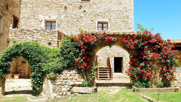 A stone building with a rustic entrance, a small wooden staircase, and a garden area with red flowers. - Rural house for exclusive rental in Girona (Cabanelles)