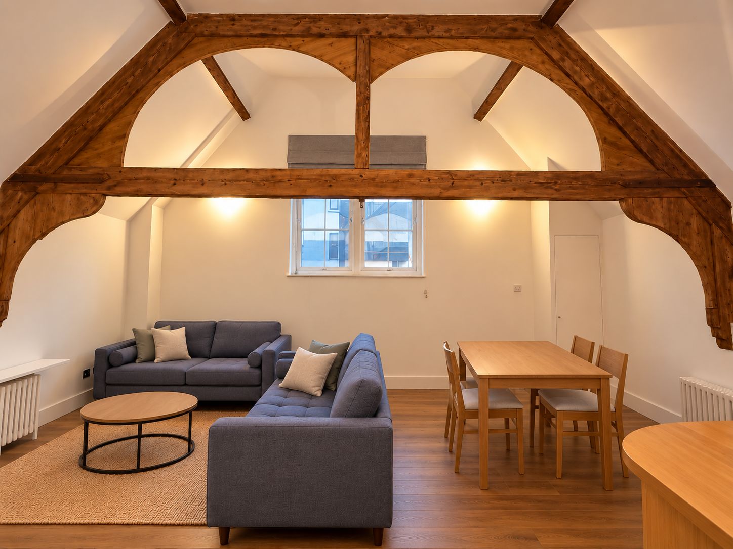A loft-style room with exposed wooden beams, a gray sofa, a wooden dining table with chairs, and a small round table.