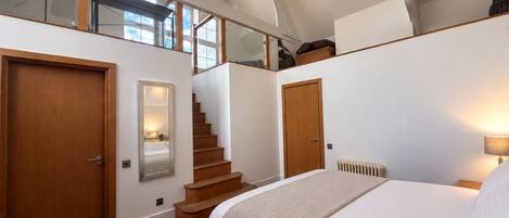 A bedroom with a large bed, a wooden staircase, and a view of the sky through the ceiling.