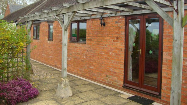 Een bakstenen gebouw met een houten pergola, een glazen deur en een kleine tuin met paarse bloemen.
