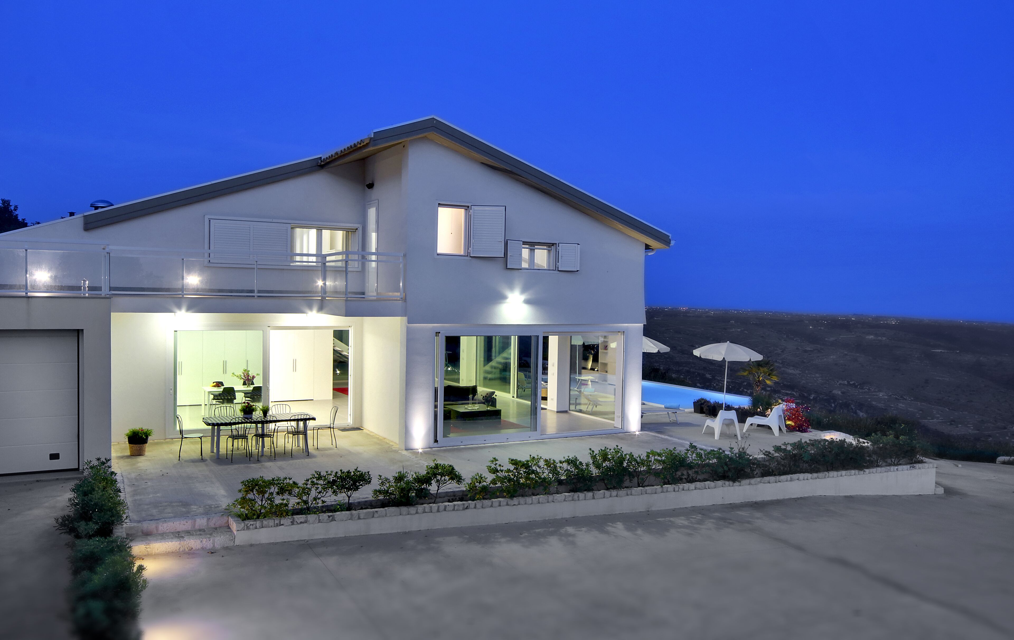 A modern two-story house with a balcony, large windows, and a view of the landscape.