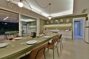 A modern dining area with a long table set for two, a kitchen in the background, and a view of the outdoors.