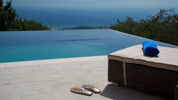 Una piscina infinita con vistas al océano, una silla de descanso con una toalla y un par de chanclas en la terraza.