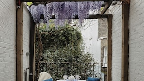 A small outdoor dining area with a checkered tablecloth, two chairs, and a white table set with plates, cups, and a teapot.
