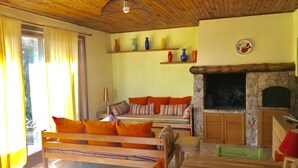 Ein gemütliches Wohnzimmer mit Holzbalkendecke, Kamin aus Stein, einer Couch mit orangenen Kissen und einem Tisch.