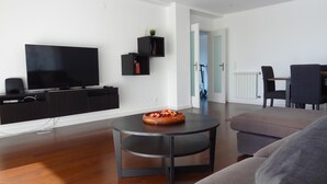 Een moderne woonkamer met een flatscreen televisie, zwarte wandplanken, een ronde salontafel met een schaal, een grijze bank en een eethoek met stoelen.