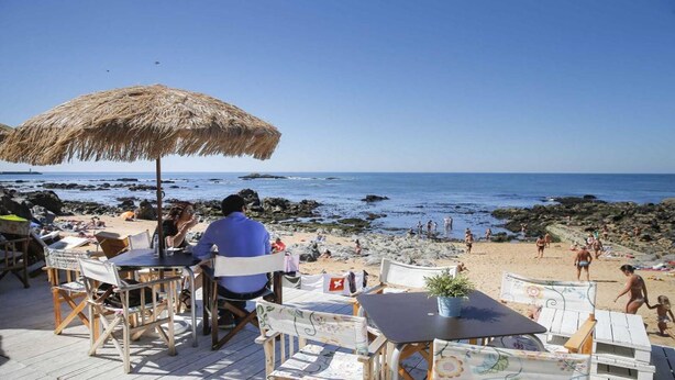 Een restaurant aan het strand met terras, een rieten parasol en uitzicht op de rotsachtige kustlijn en zee.