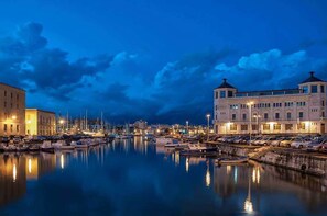 A marina with boats docked, a large building with a tower, and a calm waterfront at dusk. - Super FilinonaSuite offer at the gates of Ortigia. Fiber free and self check-in. (Siracusa)
