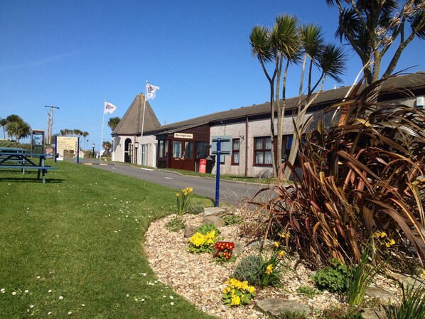 A reception building with a sign, a picnic table, and a flower bed with yellow flowers. - 3 bedroom house With Shared Use Of Pool, Tennis Courts. (, Cornwall,)