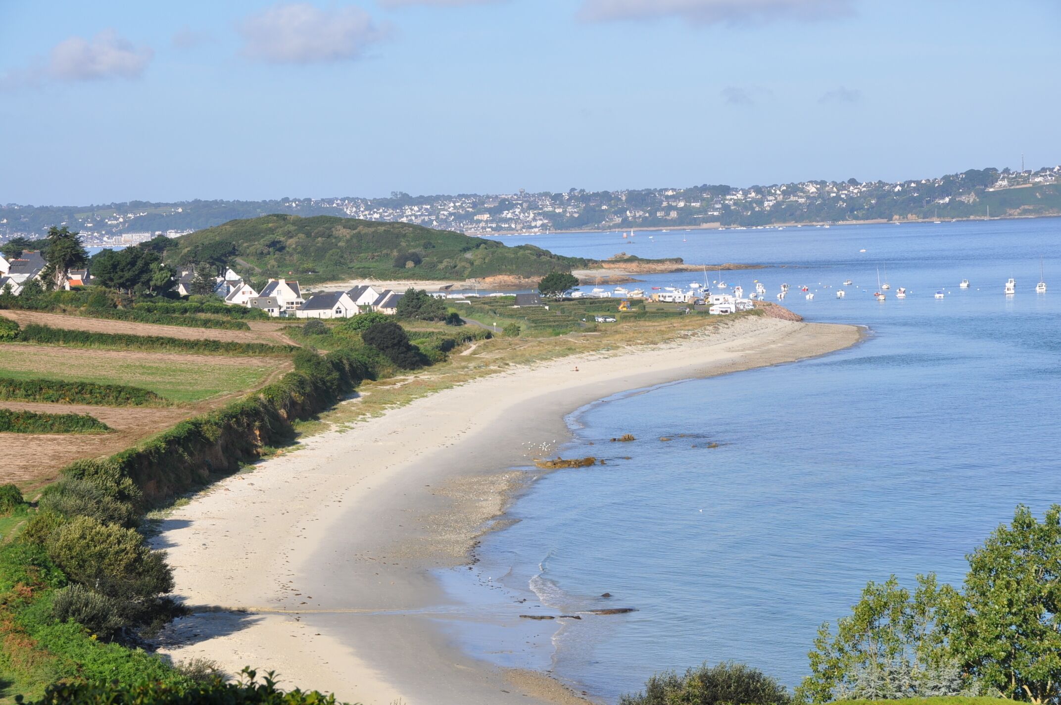 Un paisaje costero con una playa arenosa, un mar azul cristalino y una vista distante de casas y vegetación.