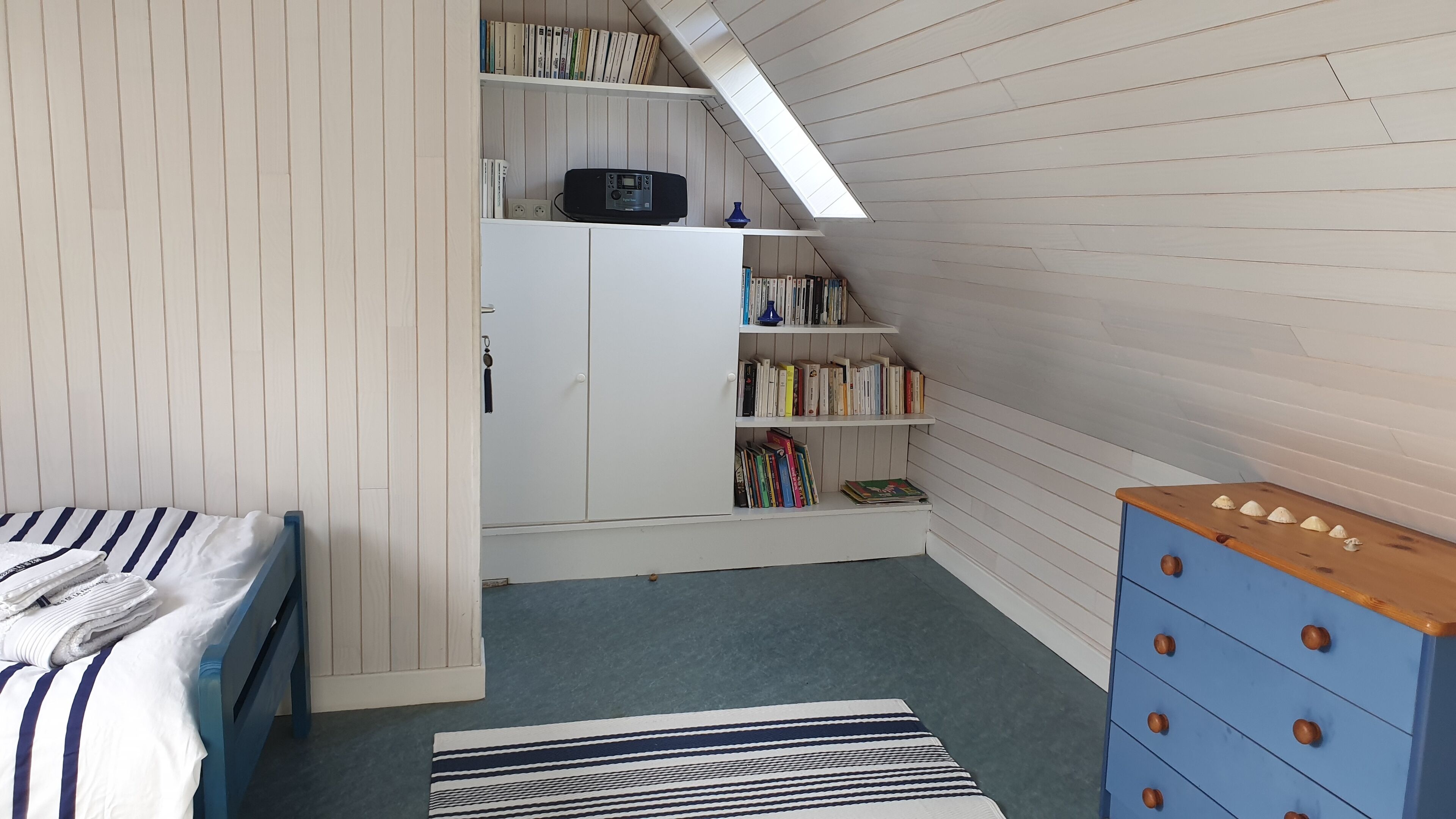 Un dormitorio con techo inclinado, una cama con ropa de cama a rayas, un aparador azul y una estantería llena de libros.