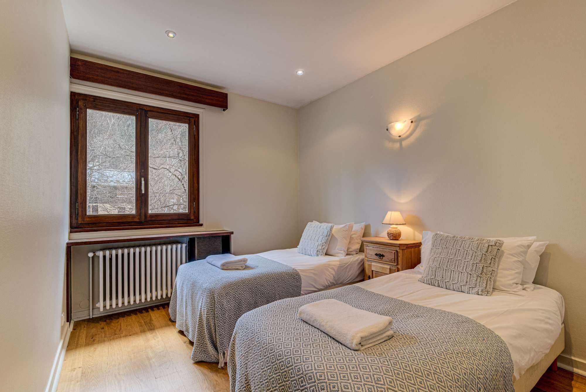 A bedroom with two beds, a radiator, a window with a view of trees, and a wall-mounted light fixture.