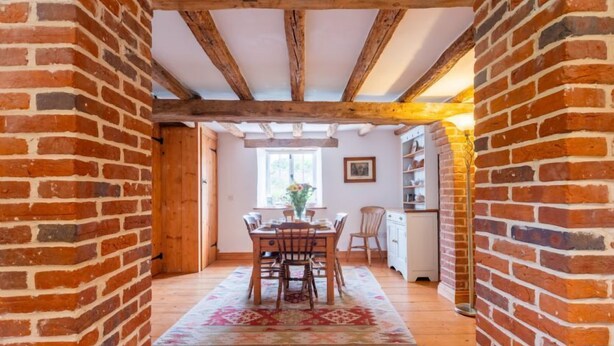 Un interior rústico con vigas de madera a la vista, pared de ladrillo, zona de comedor con mesa y sillas, y espacio de cocina con armarios y ventana.