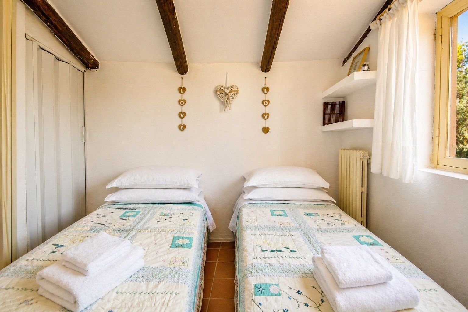 Habitación con dos camas, cada una con colcha y toalla enrollada. Vigas de madera en el techo, un adorno en la pared con corazones y una ventana con cortinas.