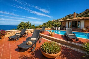 A poolside area with lounge chairs, a pool, and a view of the sea.